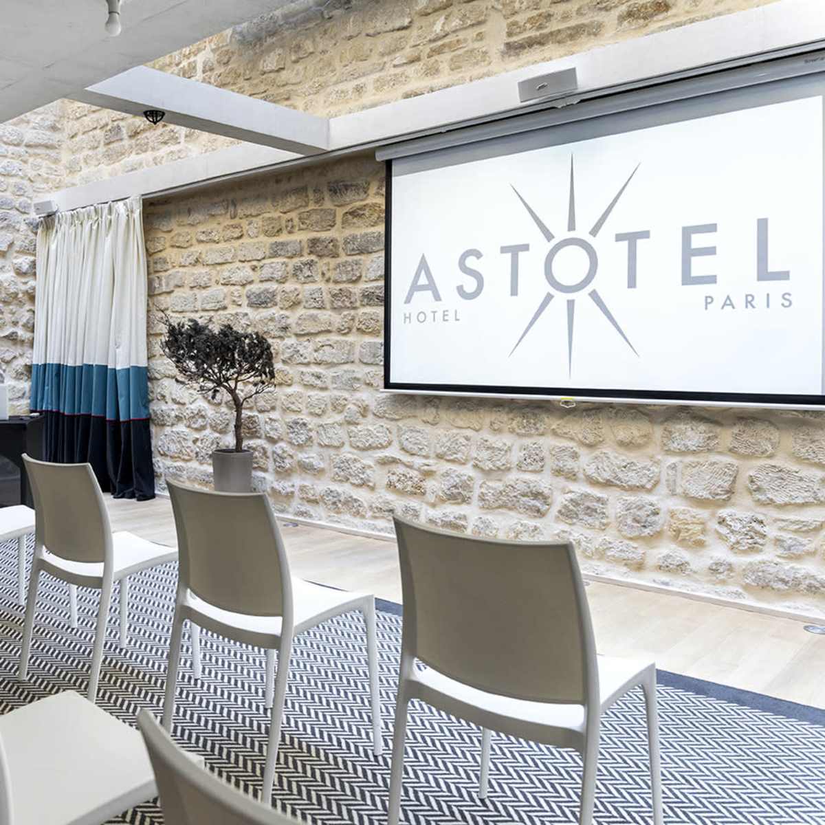 A meeting room with white chairs arranged in rows, a presentation screen displaying the "ASTOTEL PARIS" logo, and stone walls decorated with a potted plant and floral arrangements.