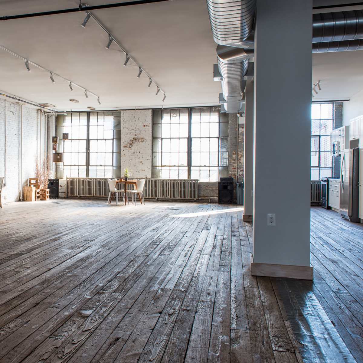 A spacious, empty room with wooden floors, large windows, and a few pieces of furniture including a table and chairs.