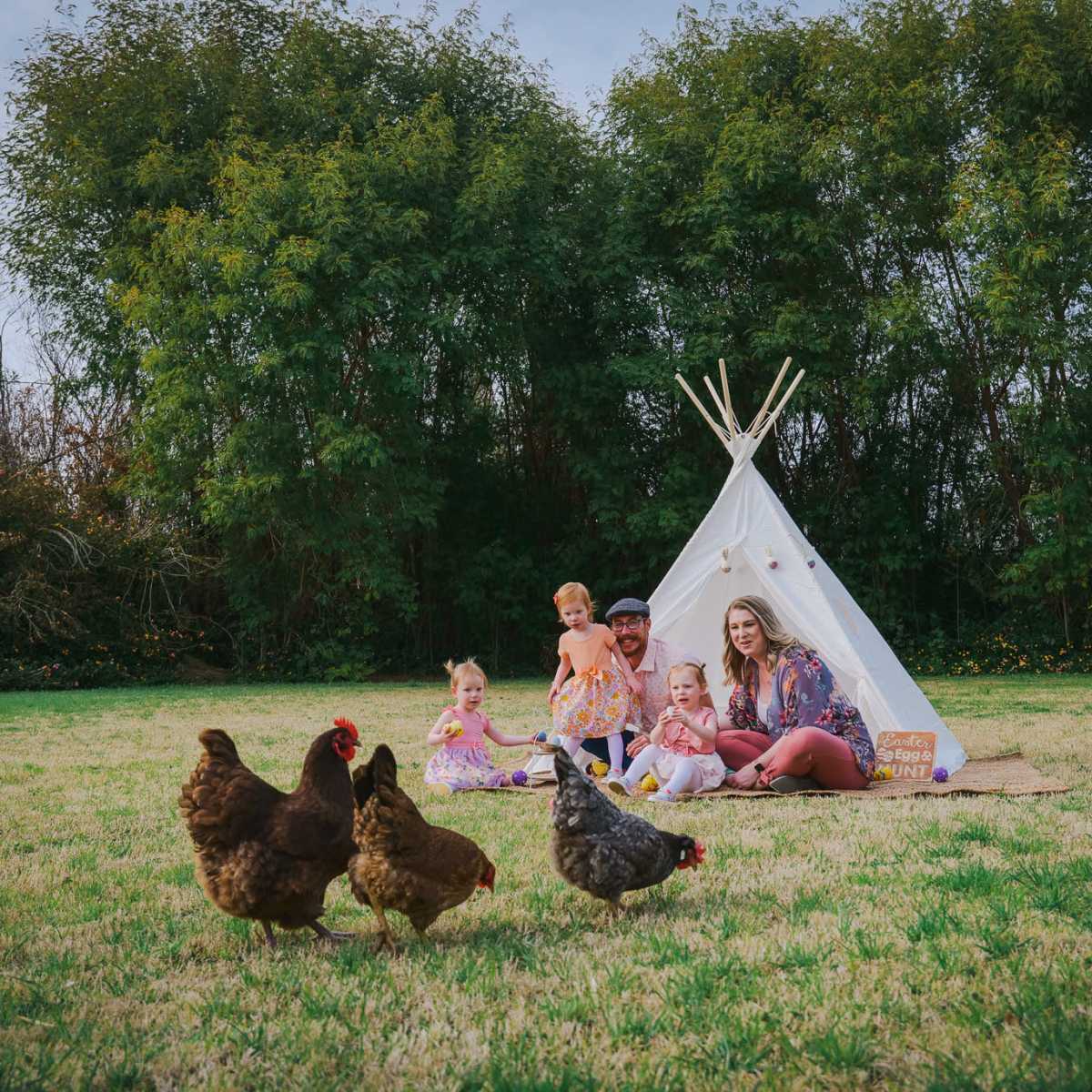 A family and several children sit on the grass in front of a teepee while chickens roam nearby.