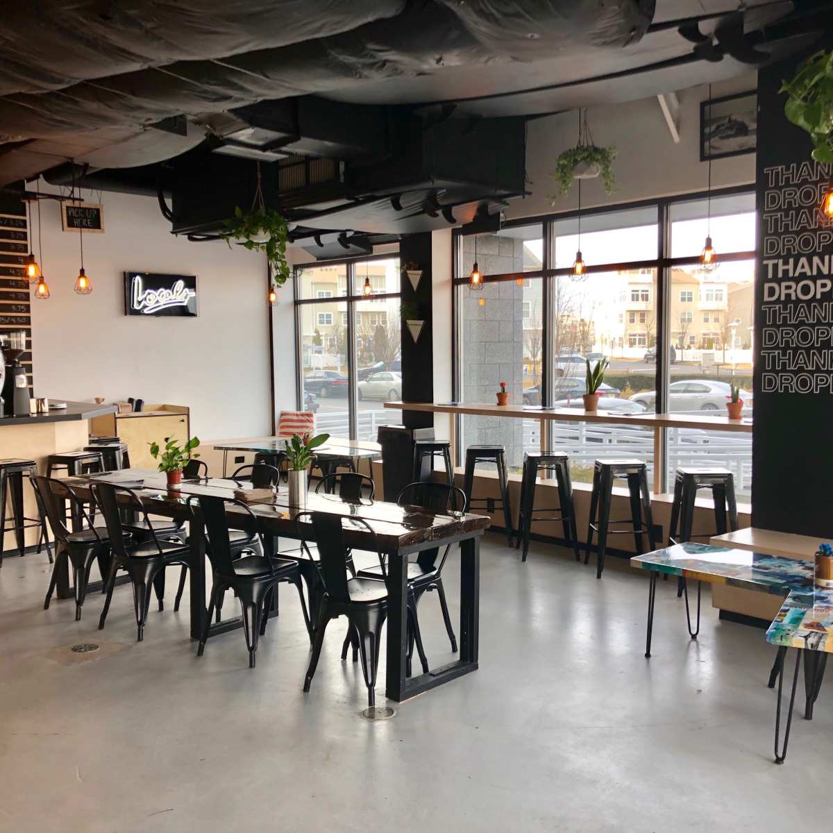 A modern café interior with wooden tables, black metal chairs, and large windows letting in natural light, featuring a coffee bar and decorative plants.