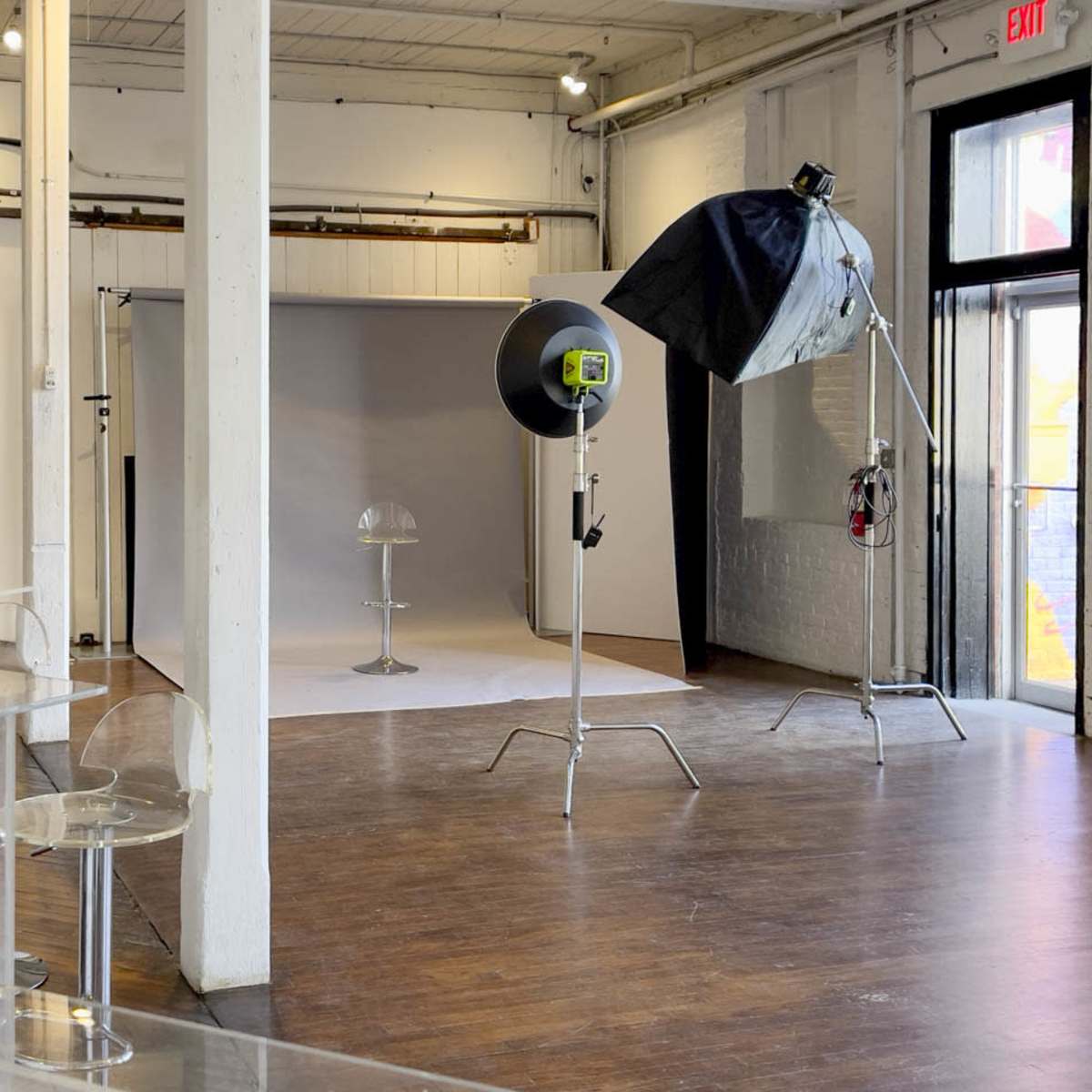 A photography studio features a white backdrop, a stool, and lighting equipment set up on a wooden floor.