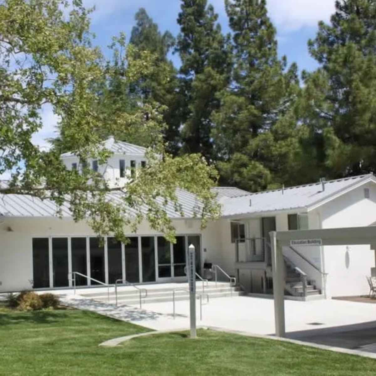 A modern building with large glass doors sits in a green landscape, surrounded by tall trees.