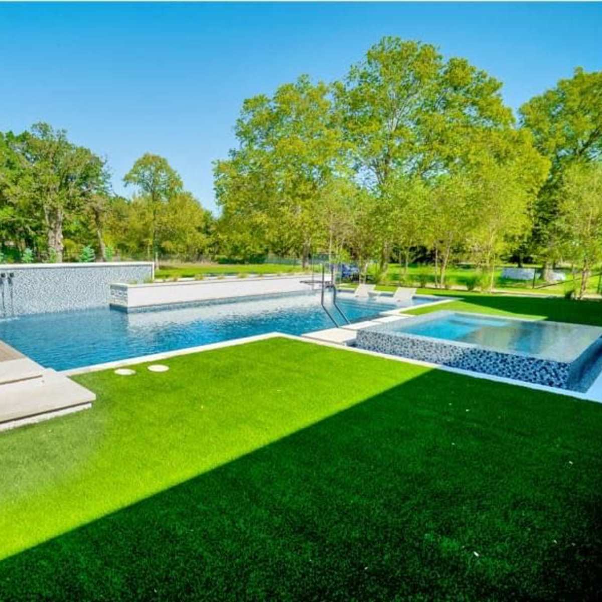 A modern swimming pool surrounded by greenery, with lounging chairs and a hot tub nearby.