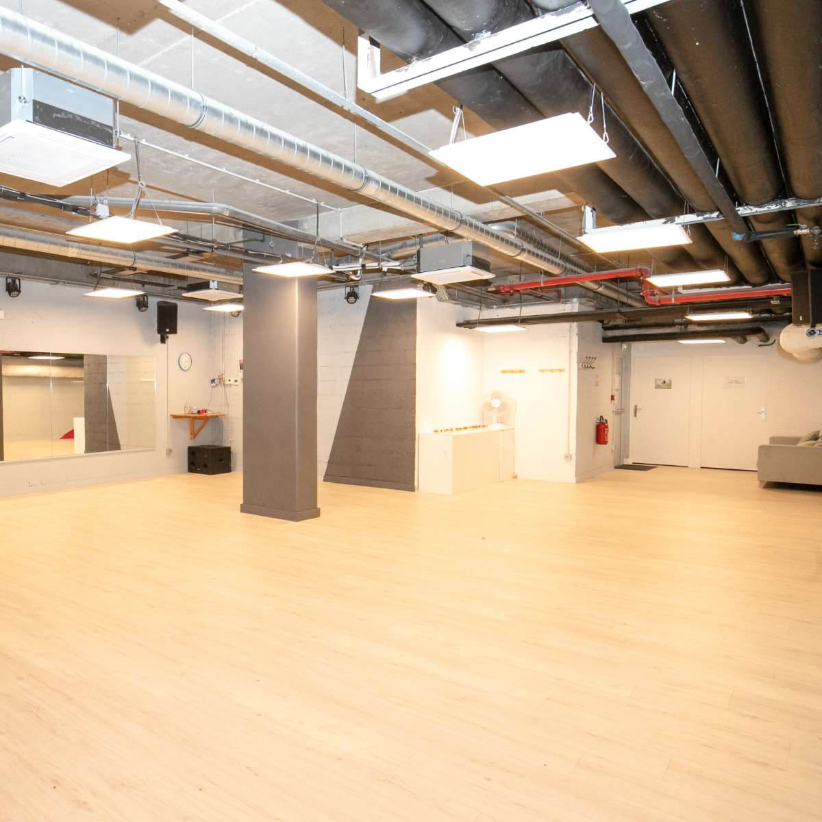 A spacious, well-lit studio with wooden flooring, mirrors on one wall, and exposed piping on the ceiling.
