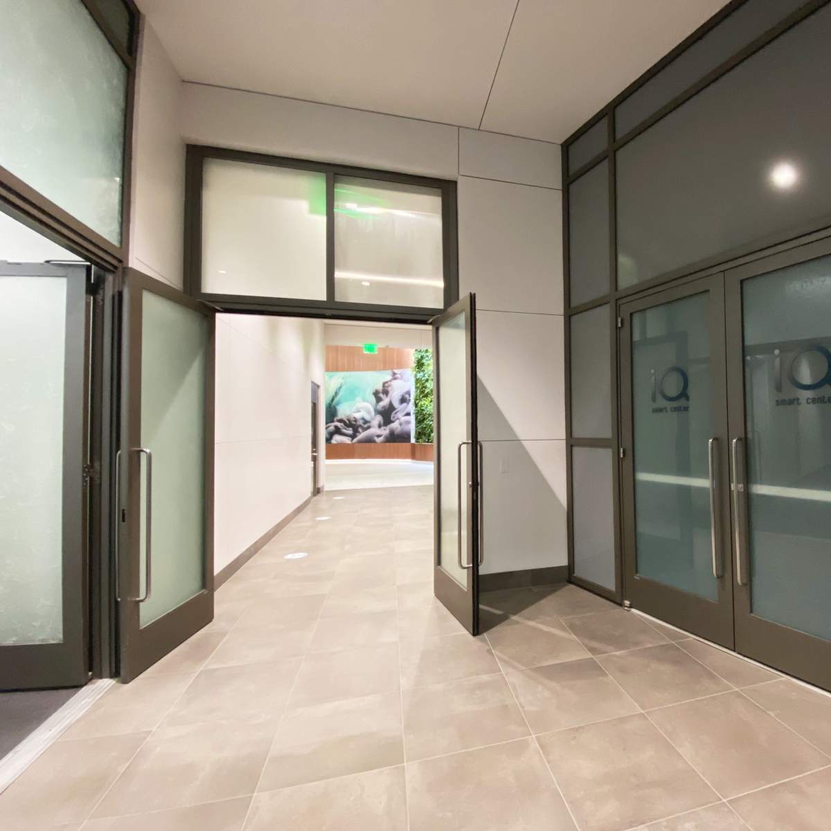 A hallway with glass doors on either side and a tiled floor, leading to another room with greenery visible in the background.