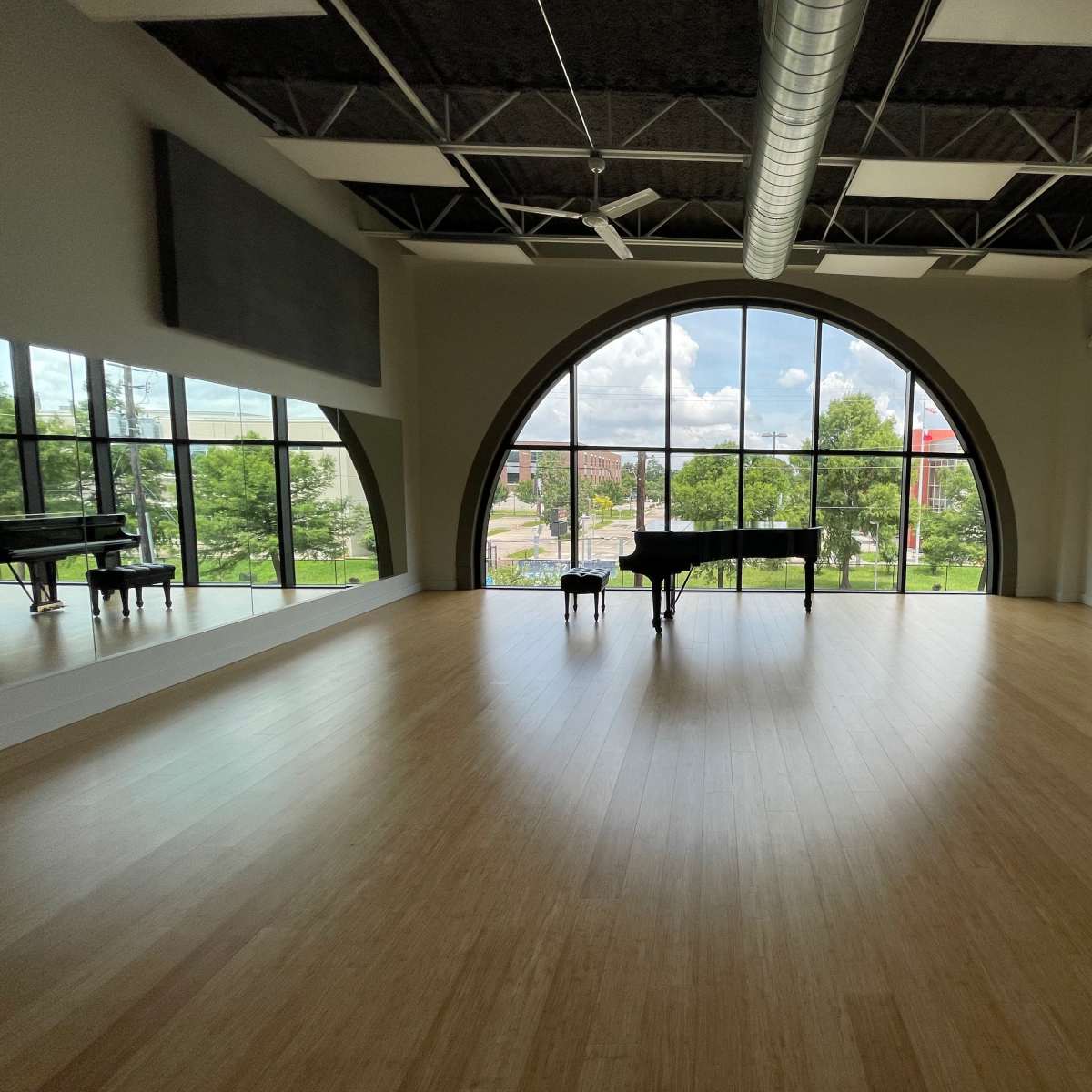 A spacious music studio features a grand piano positioned in front of a large arched window, overlooking a tree-lined street.