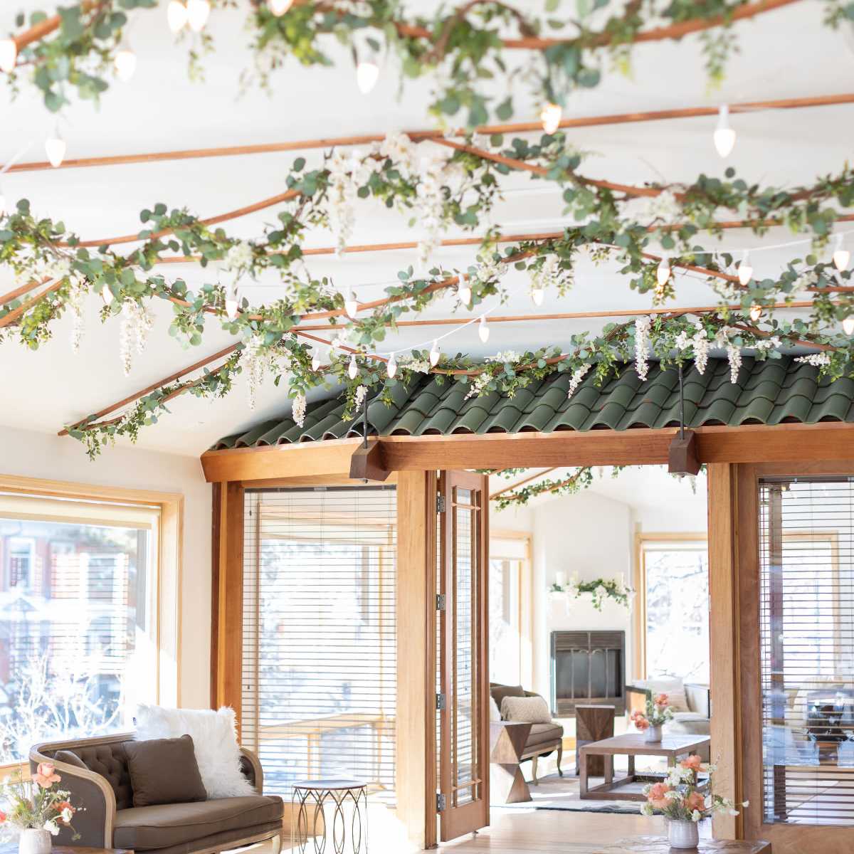 A cozy room features a wooden ceiling adorned with greenery and string lights, with furniture arranged around a central area near large windows.