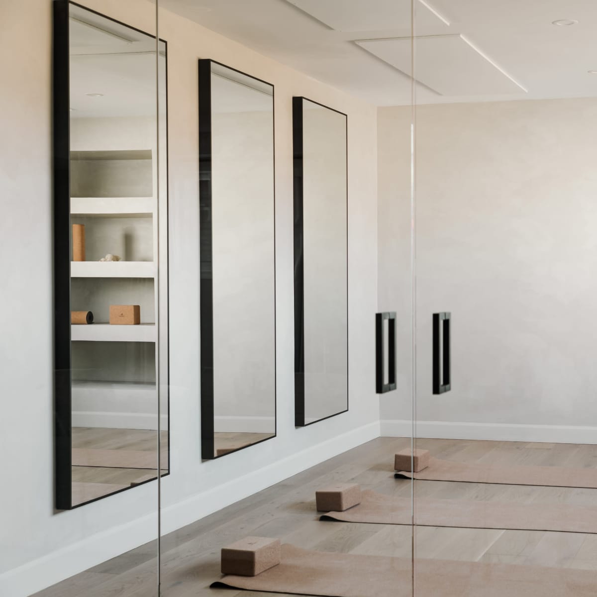 A minimalist room with three large mirrors on the wall, two yoga mats on the floor, and shelves displaying a few objects.