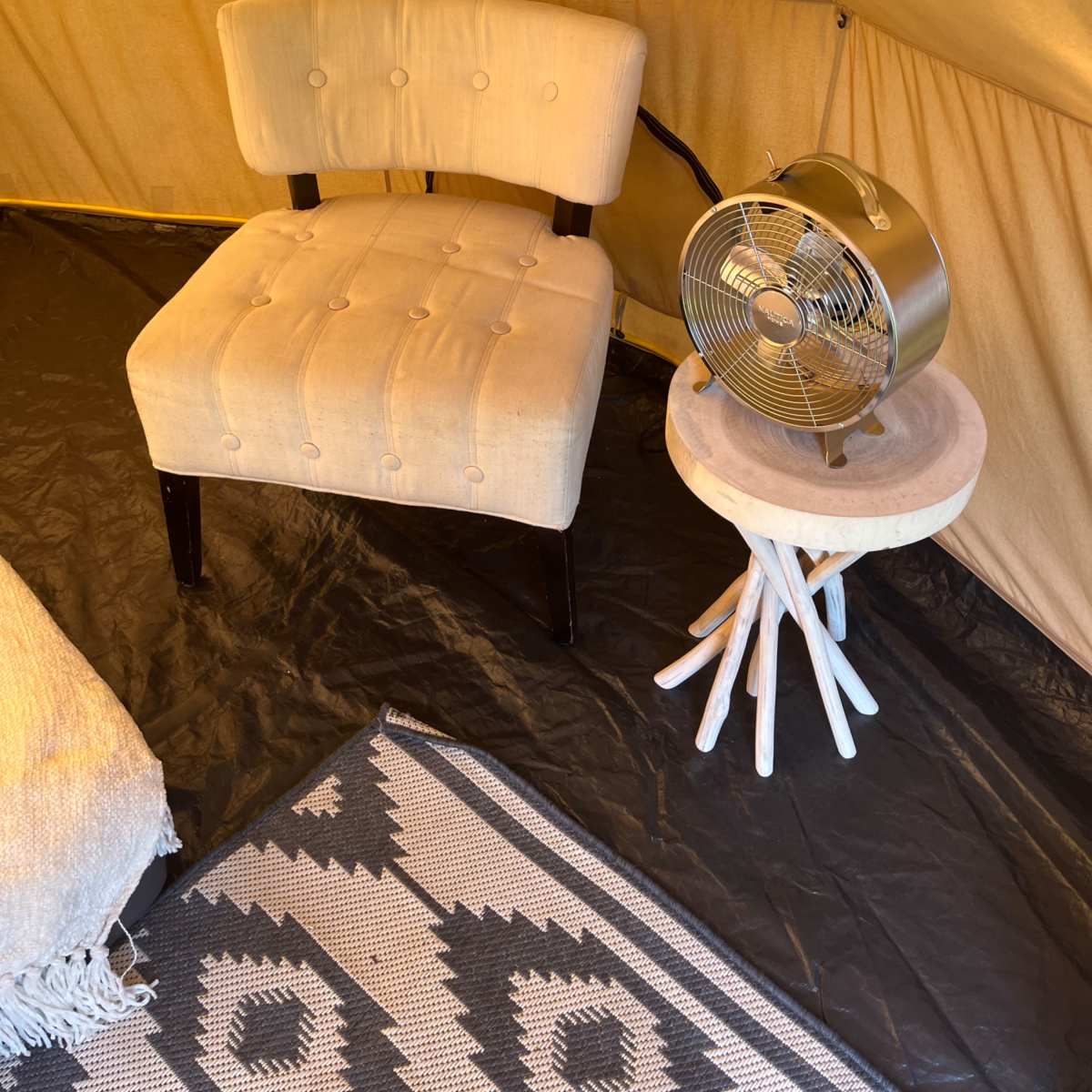 An interior of a tent featuring a beige upholstered chair, a small wooden side table with a fan, and a patterned rug on a black floor.