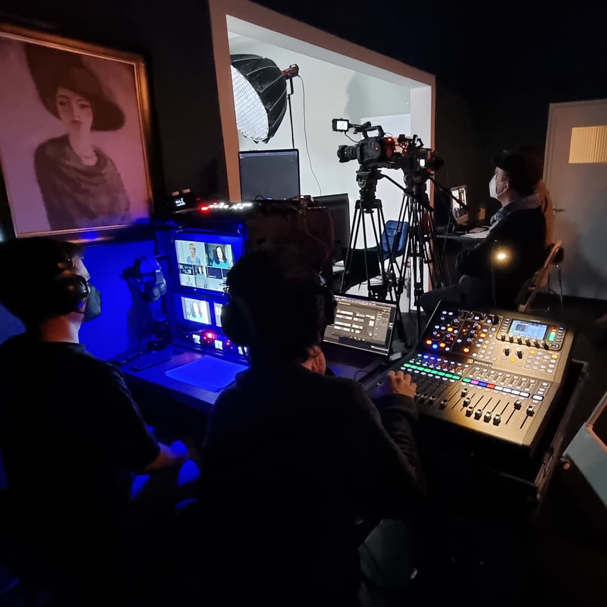 A film or video production setup, featuring multiple cameras and production equipment in a dimly lit room with a portrait hanging on the wall.