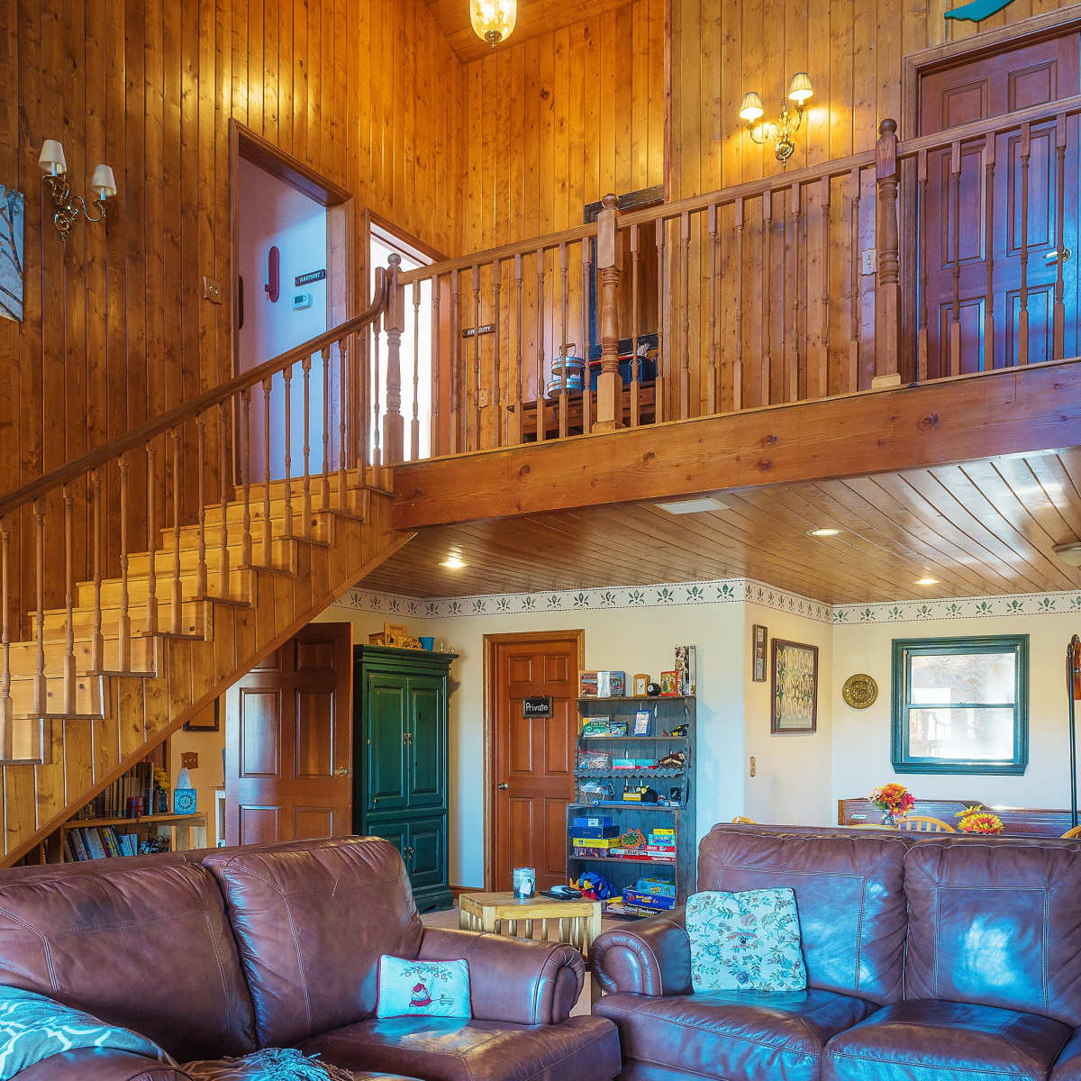 A wooden-paneled living room with two brown leather sofas, a staircase leading to an upper level, and various doors and decorations along the walls.