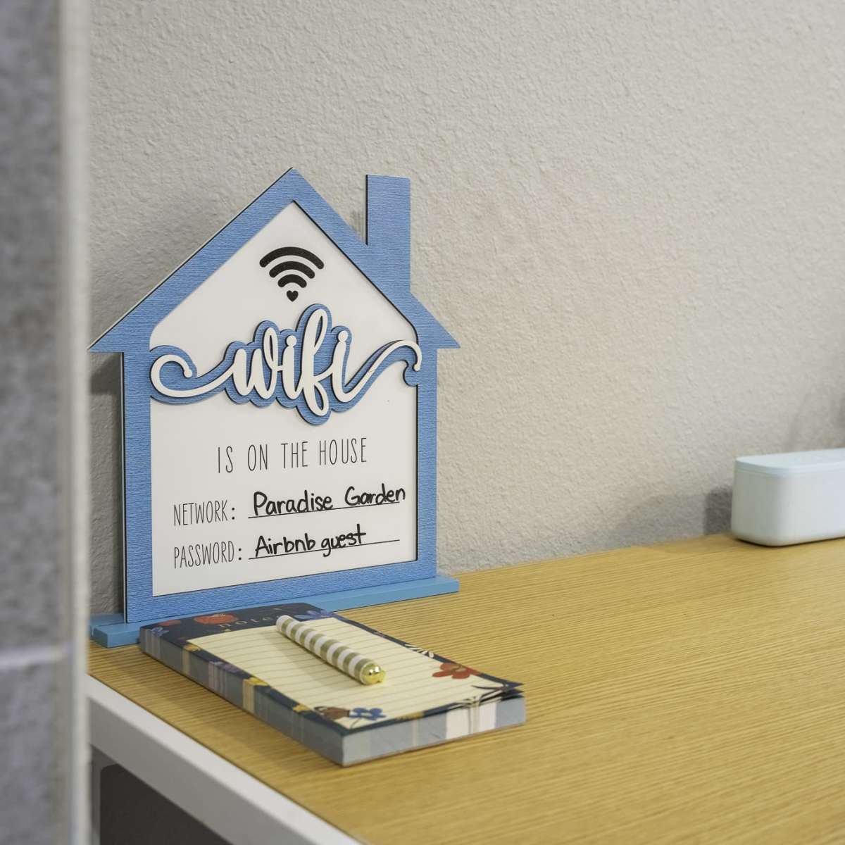 A blue and white sign displaying Wi-Fi information sits on a wooden table beside a small plant and a speaker.
