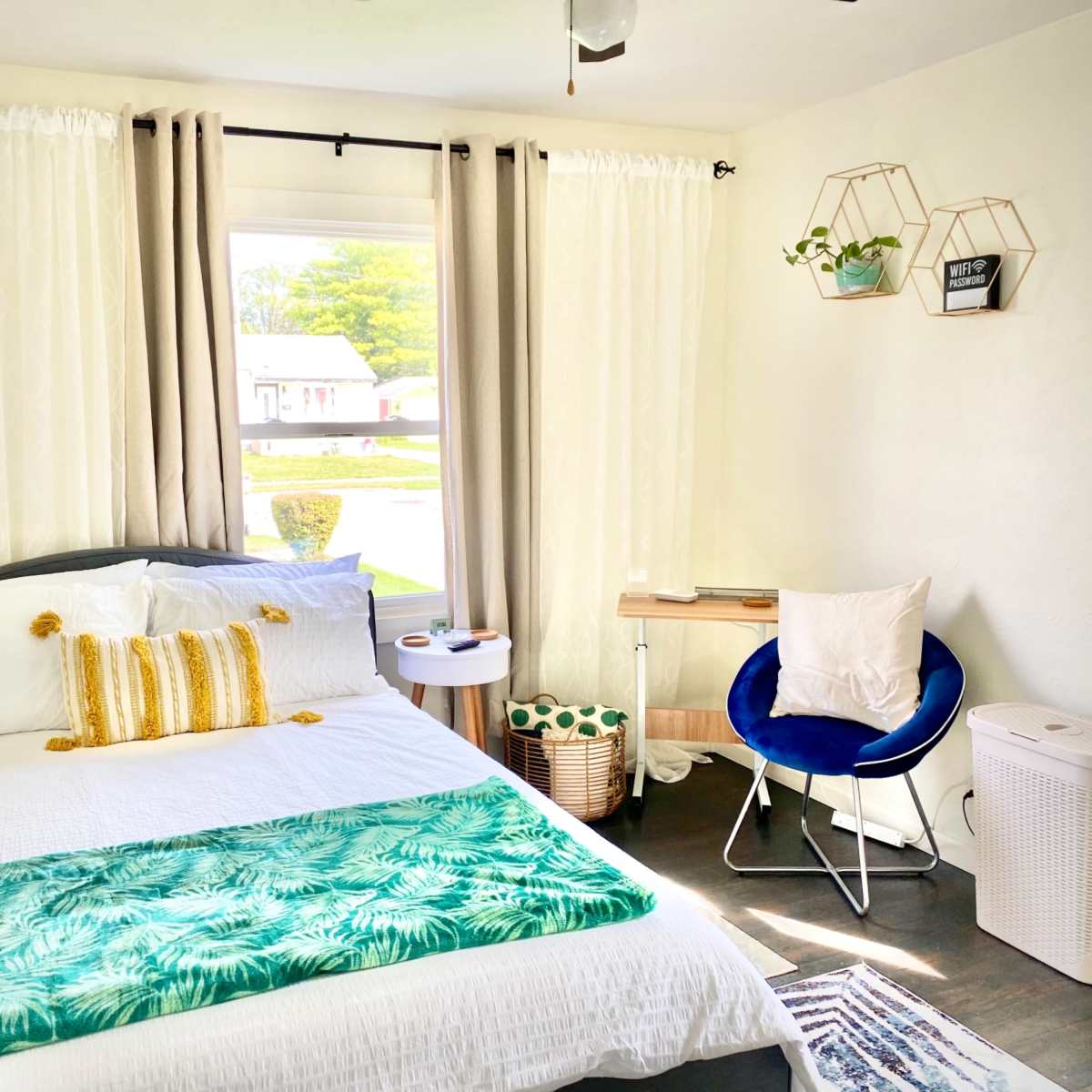 A cozy bedroom featuring a double bed with white linens, a small table, a blue chair, and large windows with light curtains.