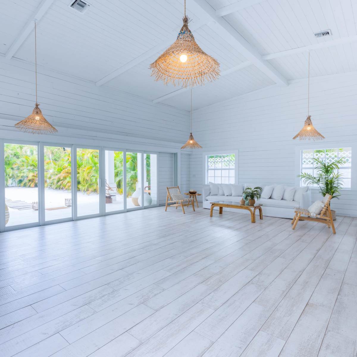 A bright, open interior space with white walls and wooden flooring, featuring a seating area with a light-colored couch and large windows that offer a view of a tropical outdoor scene.