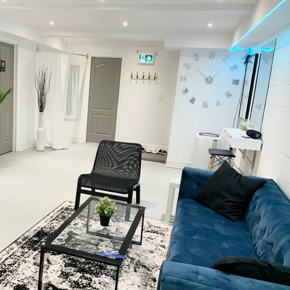 A minimalist, well-lit room with a blue sofa, a glass coffee table, a black chair, a decorative plant, and a clock on the wall.