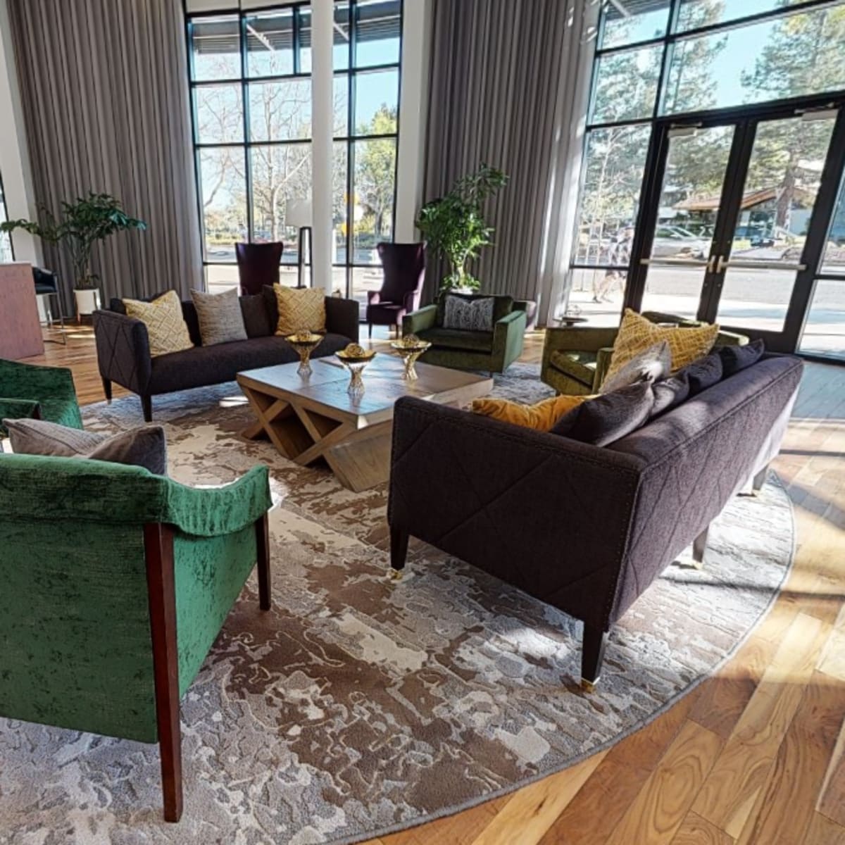 A spacious lounge area features a round rug surrounded by green and brown upholstered chairs and a central coffee table, with large windows providing natural light.