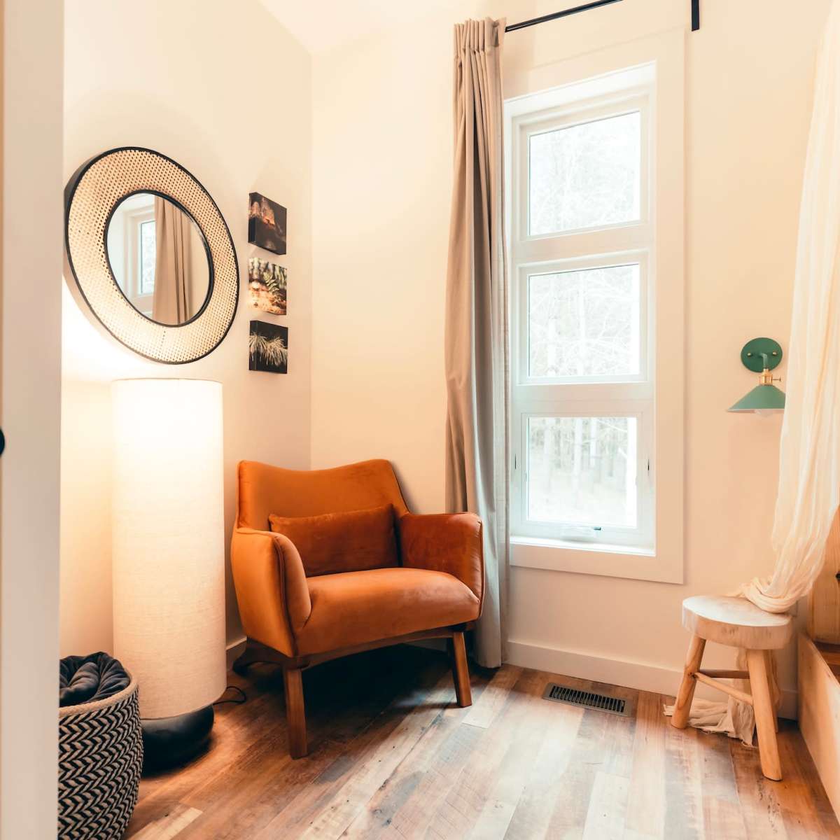 A cozy bedroom featuring a brown armchair, a tall lamp, and a window with light curtains, along with natural wood flooring.