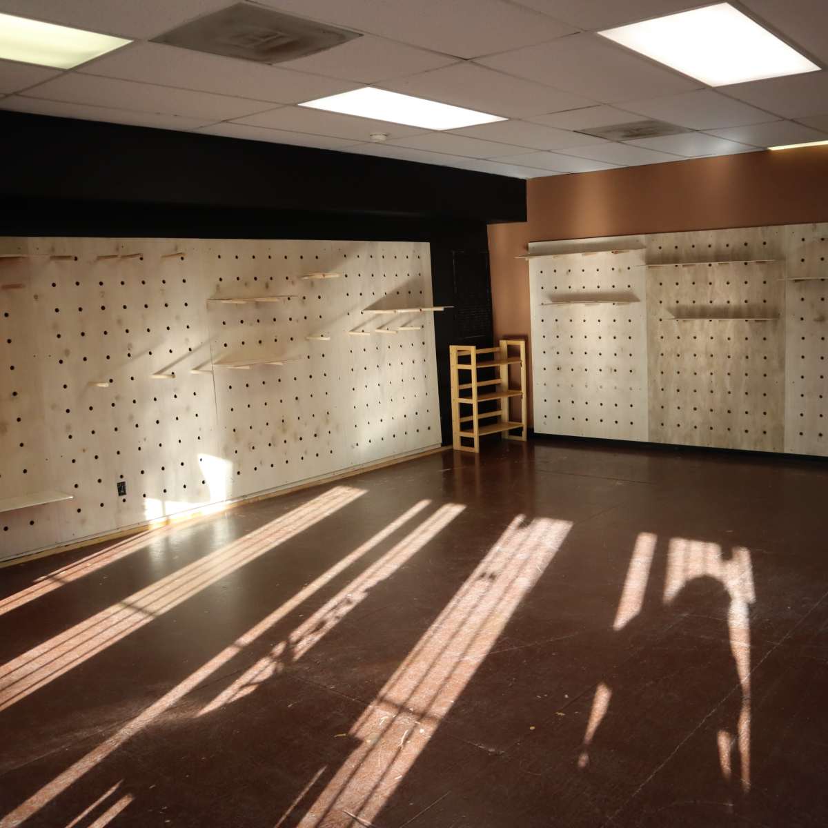 An empty room with pegboard walls and wooden furniture, illuminated by sunlight casting shadows on the floor.