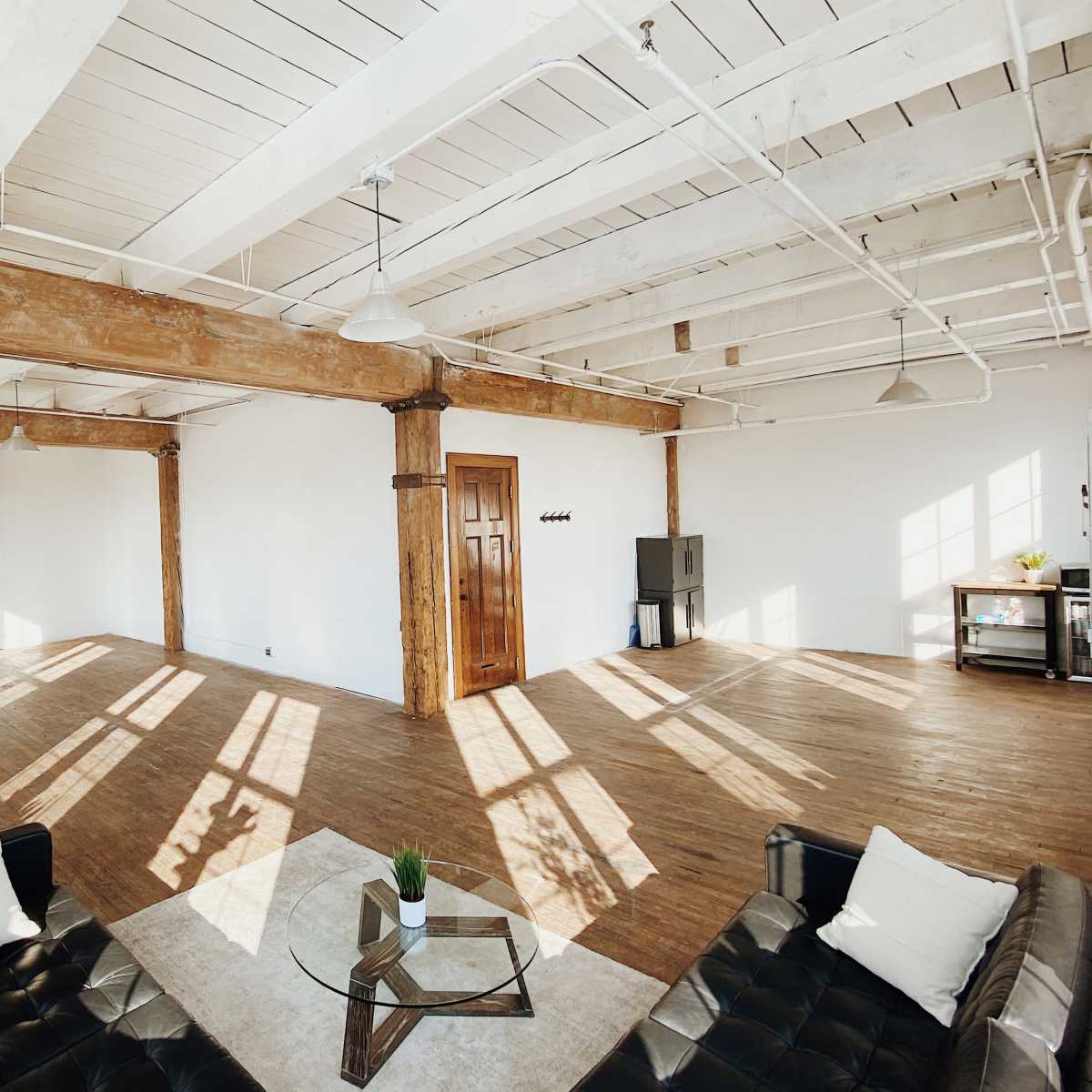 A spacious, well-lit interior with wooden beams, a dark sofa, a coffee table, and large windows casting shadows on the floor.