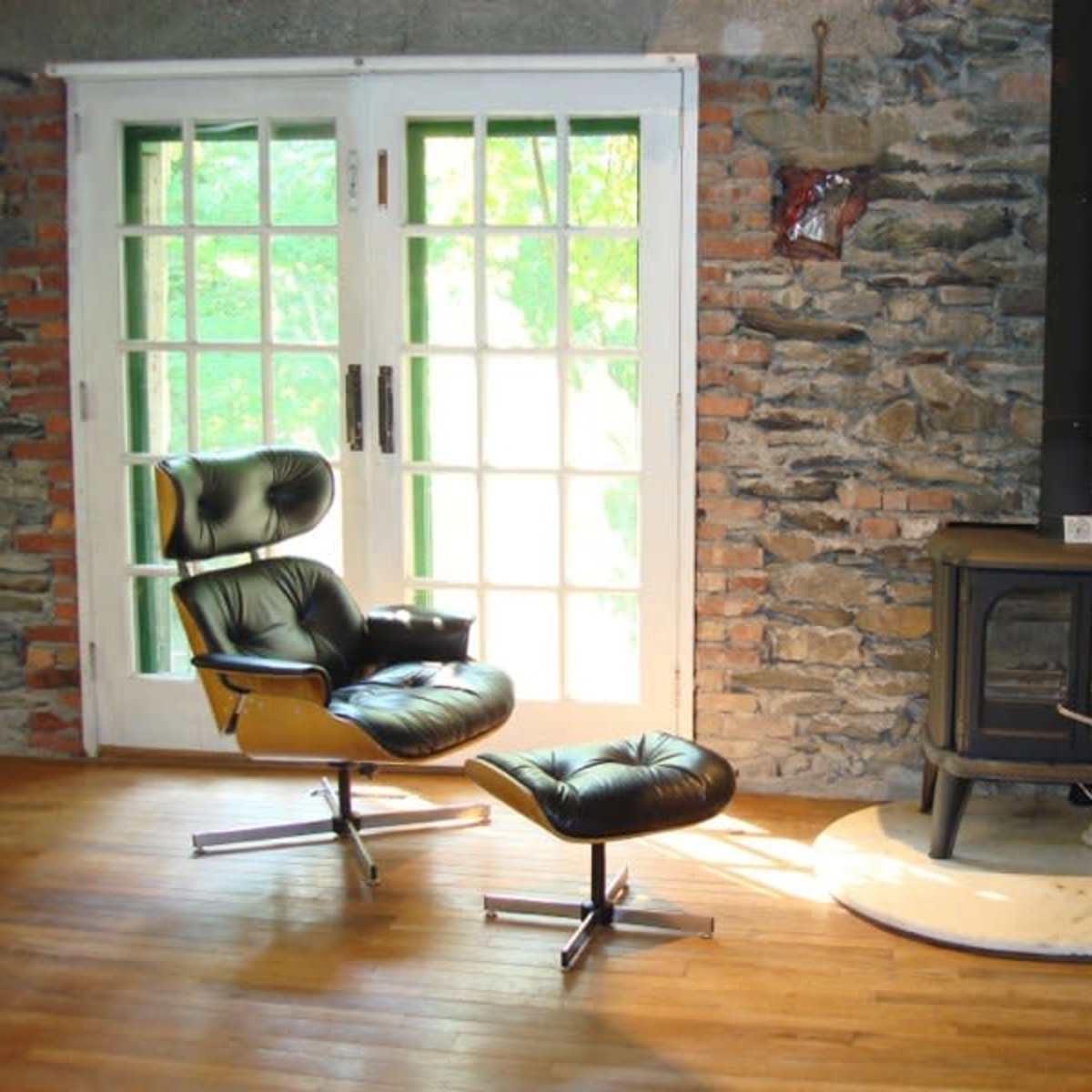 A black leather chair and ottoman sit beside a black metal wood stove on a marble pedestal, with large windows revealing a stone wall backdrop.