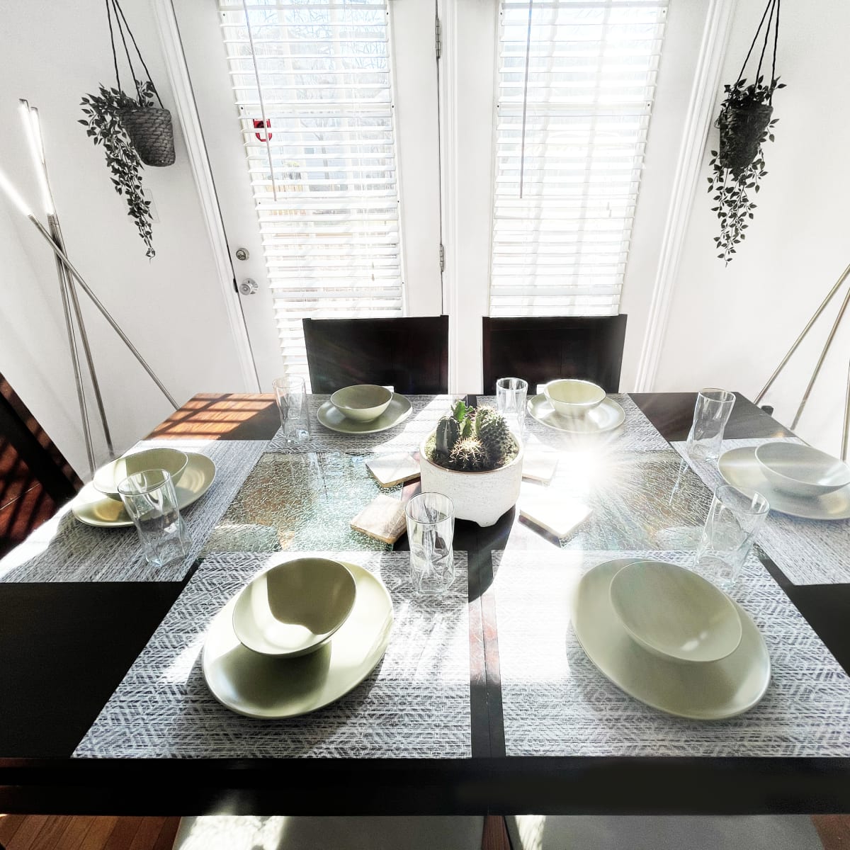 A dining table is set with green plates and glasses, surrounded by bright natural light filtering through window shutters.