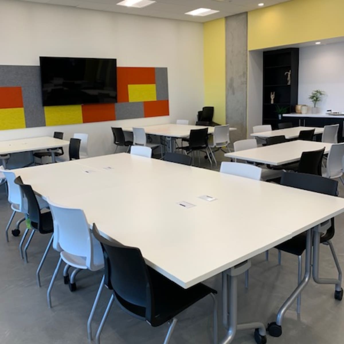 A modern conference room is arranged with multiple white tables and chairs, featuring a large television screen and colorful wall panels.