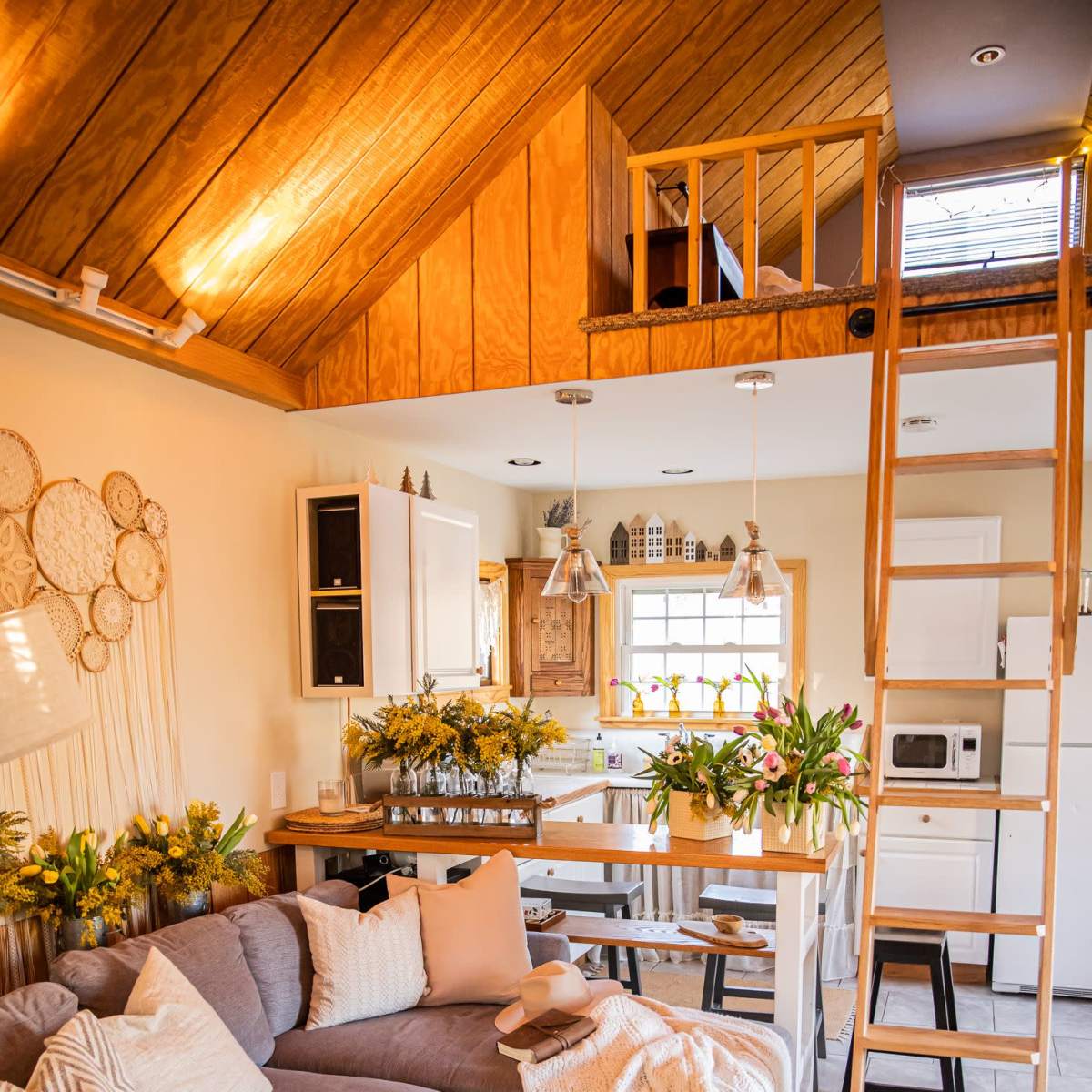 A cozy living space features a gray sectional sofa, a wooden dining table adorned with flowers, and a ladder leading to a loft area.