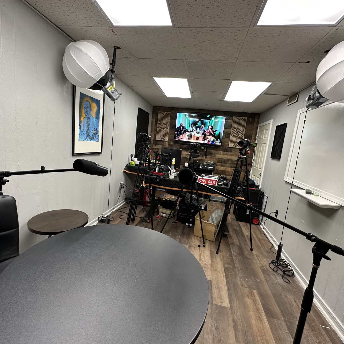 A recording studio with two microphones positioned at a round table, cameras set up in the background, and a television displaying a video on a wall.