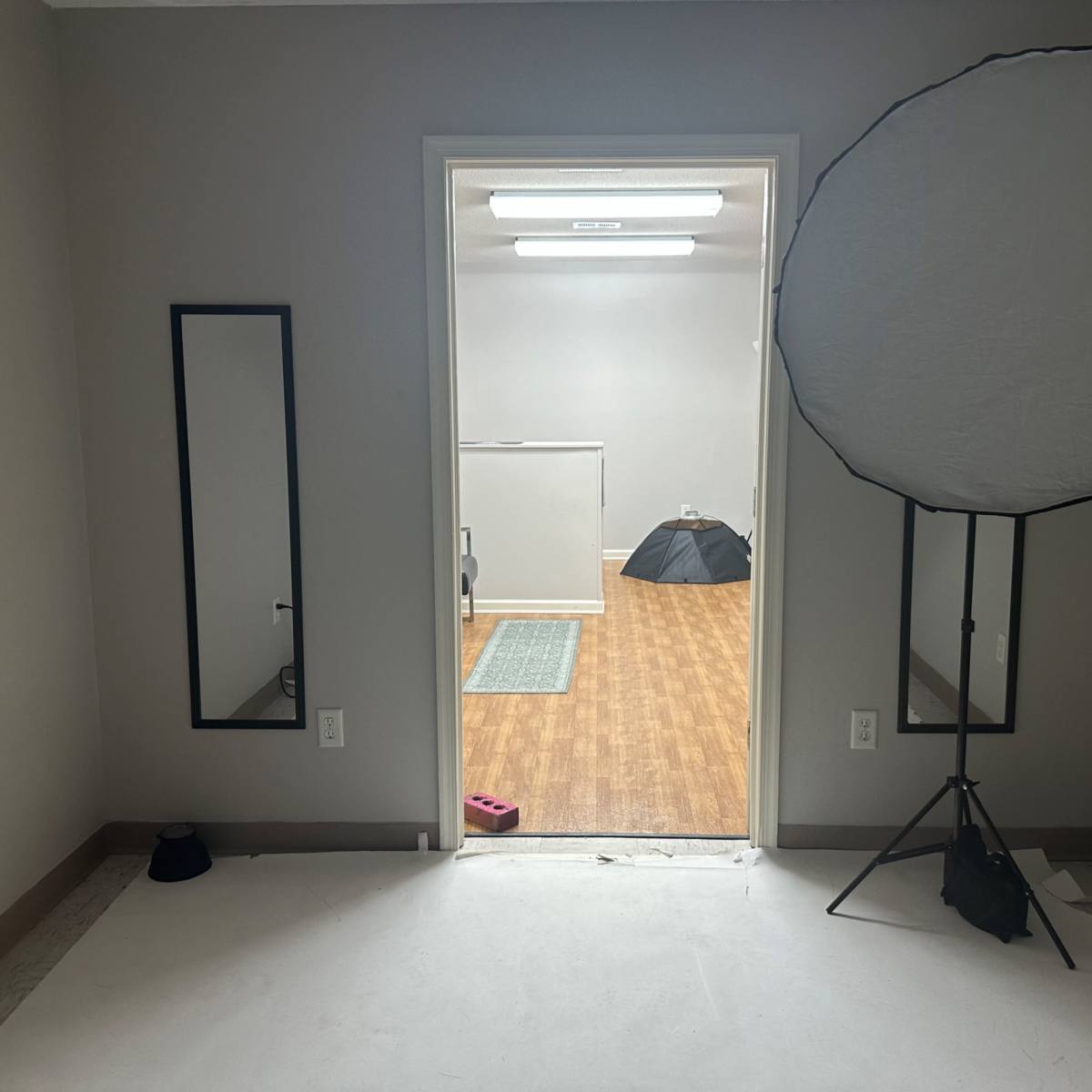 A blank room with a doorway leading into another space, featuring a mirror on the left and a large circular reflector on a stand to the right.