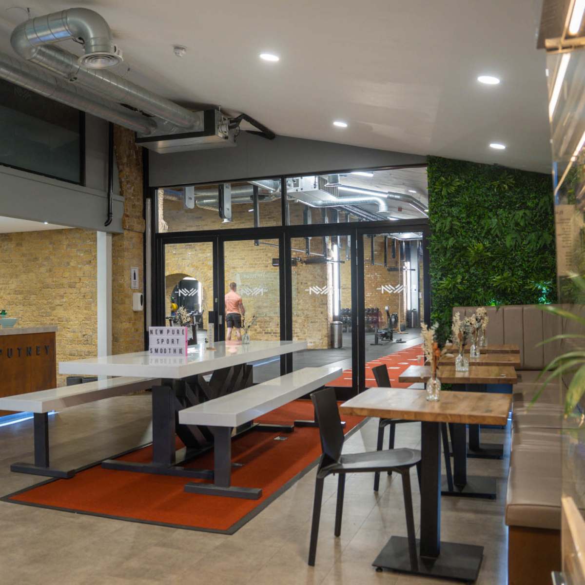 A modern, open-plan café with a reception area, large communal tables, and greenery accenting the walls.
