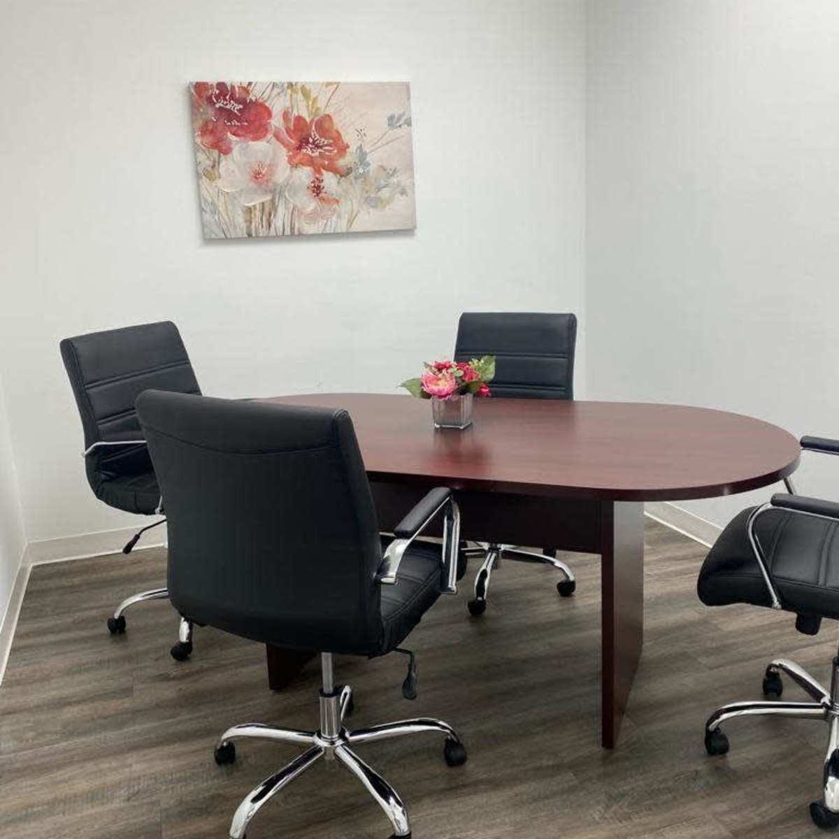 A small meeting room features a round wooden table surrounded by four black swivel chairs, with a flower arrangement in the center and a floral painting on the wall.