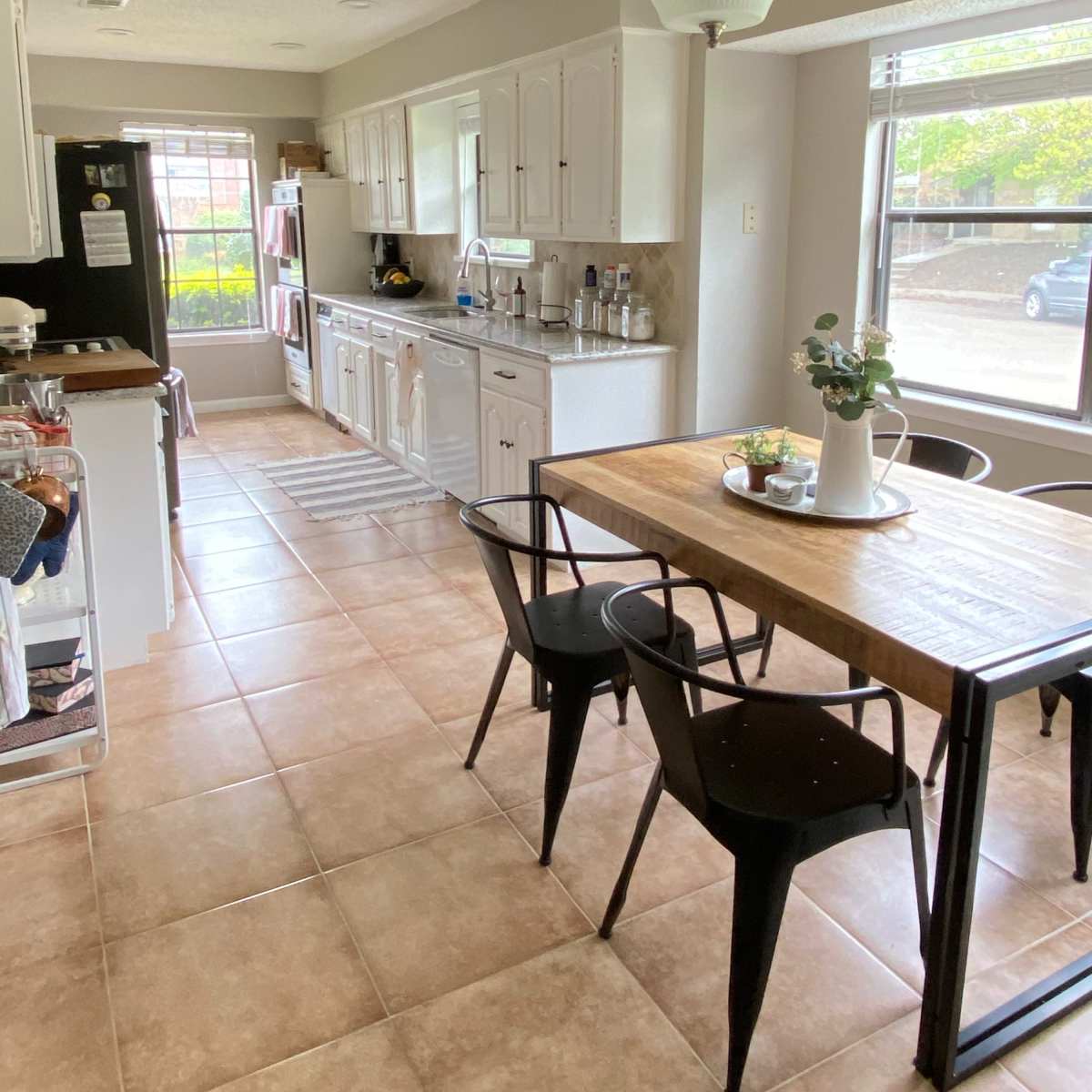 A modern kitchen with white cabinets, a wooden dining table, and a view of a patio through large windows.