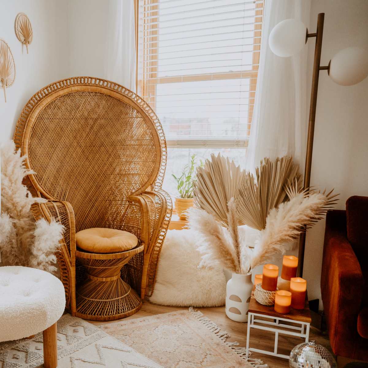 A cozy corner of a room featuring a large wicker chair, a round ottoman, and decorative plants, illuminated by natural light from a window.