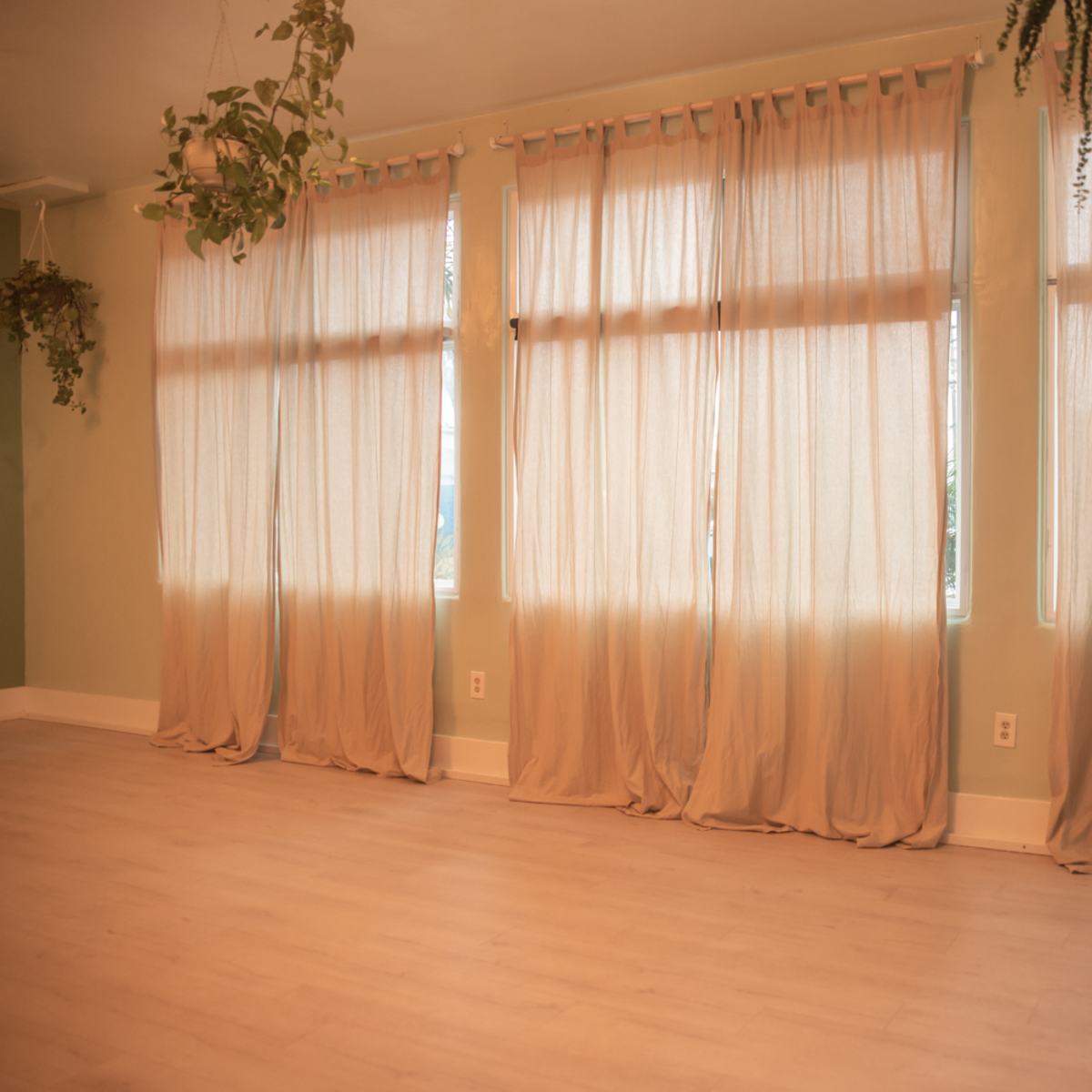 A room with large, light-colored curtains hanging in front of three windows, and plants suspended from the ceiling.