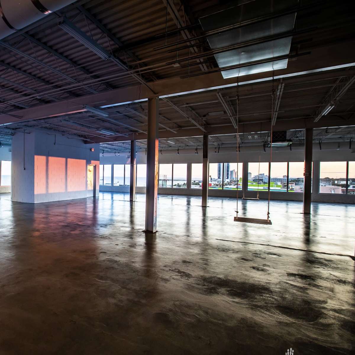 A spacious, empty room with large windows overlooking a body of water and a cityscape, illuminated by soft evening light.