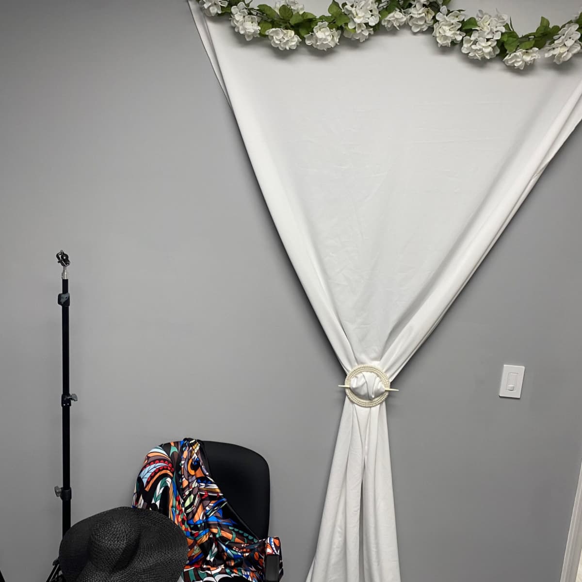 A gray wall adorned with a white drape and artificial flowers, next to a chair and a few stands.