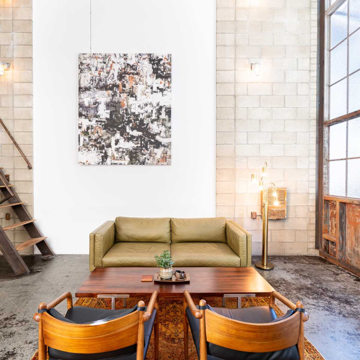 A modern living space with a green sofa, a wooden coffee table, and a large abstract painting on a white wall, alongside a staircase and large windows.