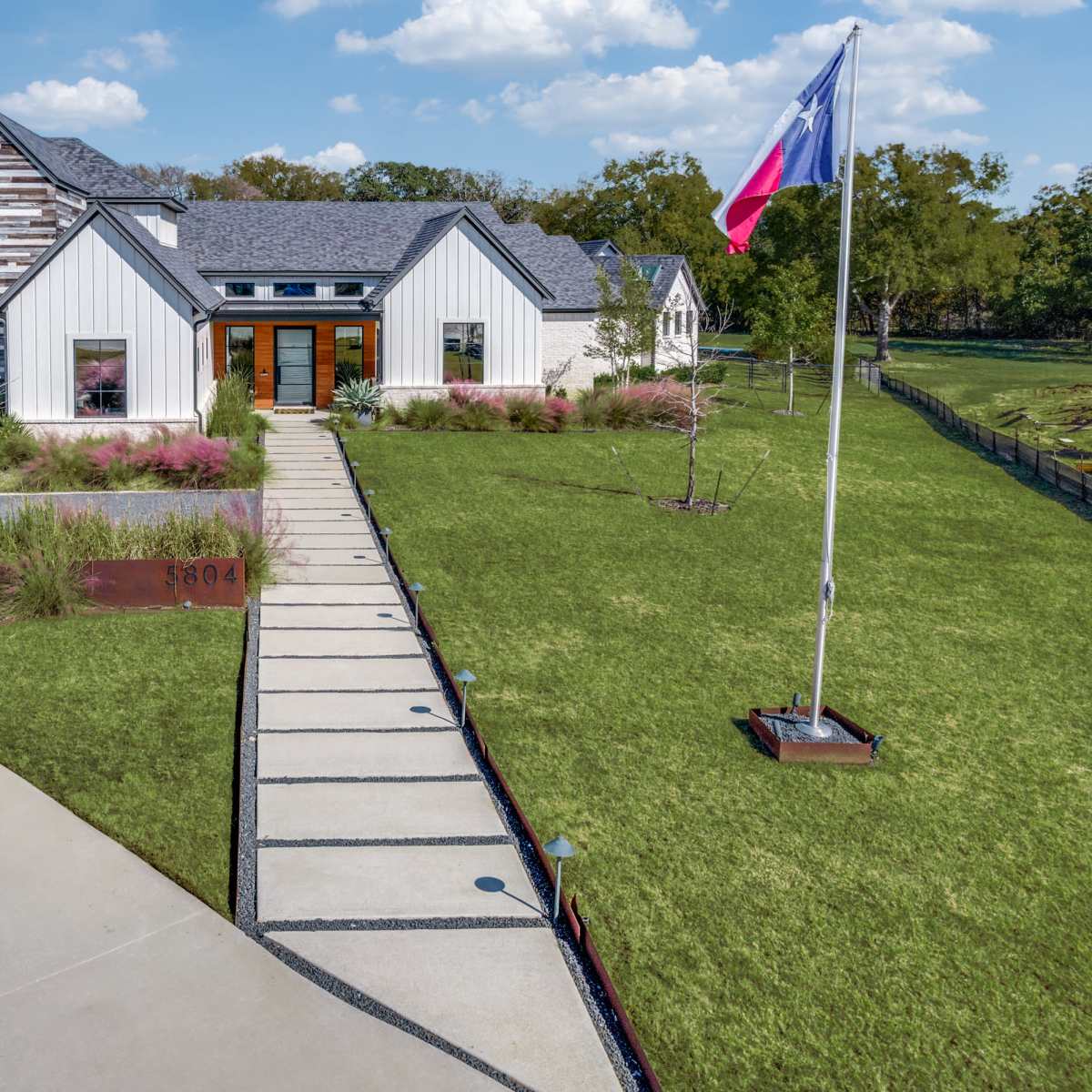 A modern house with a mix of white and gray siding, featuring large front windows and a Texas flag, is set against a landscaped lawn with a concrete path leading to the entrance.