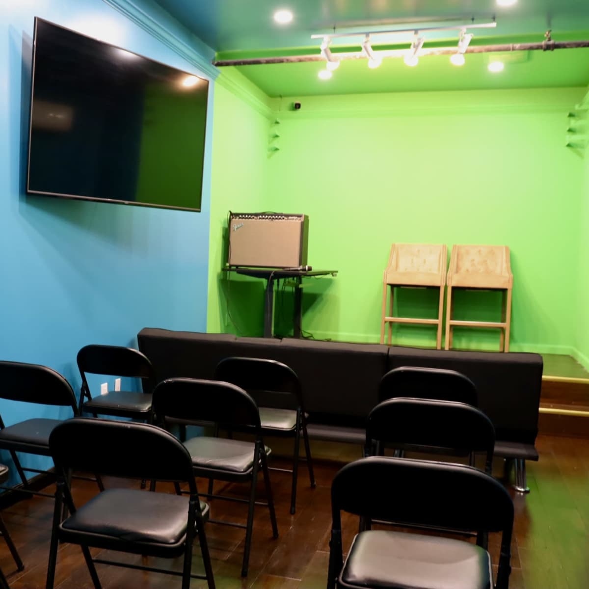 A small stage area with a blue wall, a green backdrop, an amplifier on a table, and several black chairs arranged in front.