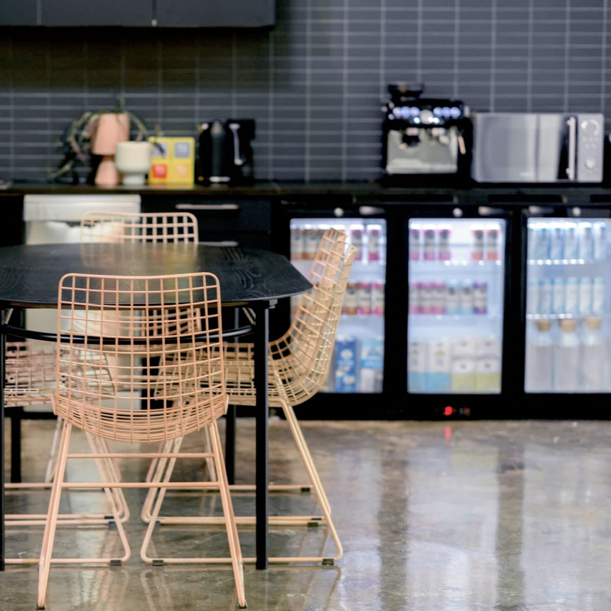 A black table with four wireframe chairs is set in a modern kitchen with dark cabinetry and stainless steel appliances.