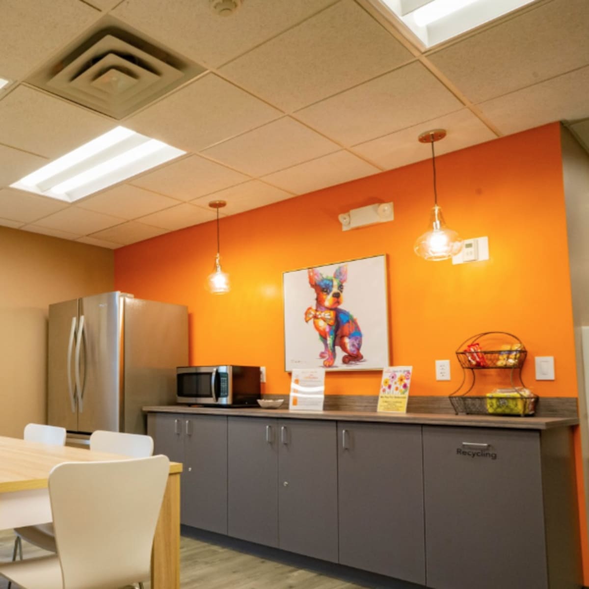 A modern break room with a wooden table, white chairs, a refrigerator, a microwave, and colorful artwork on the walls.