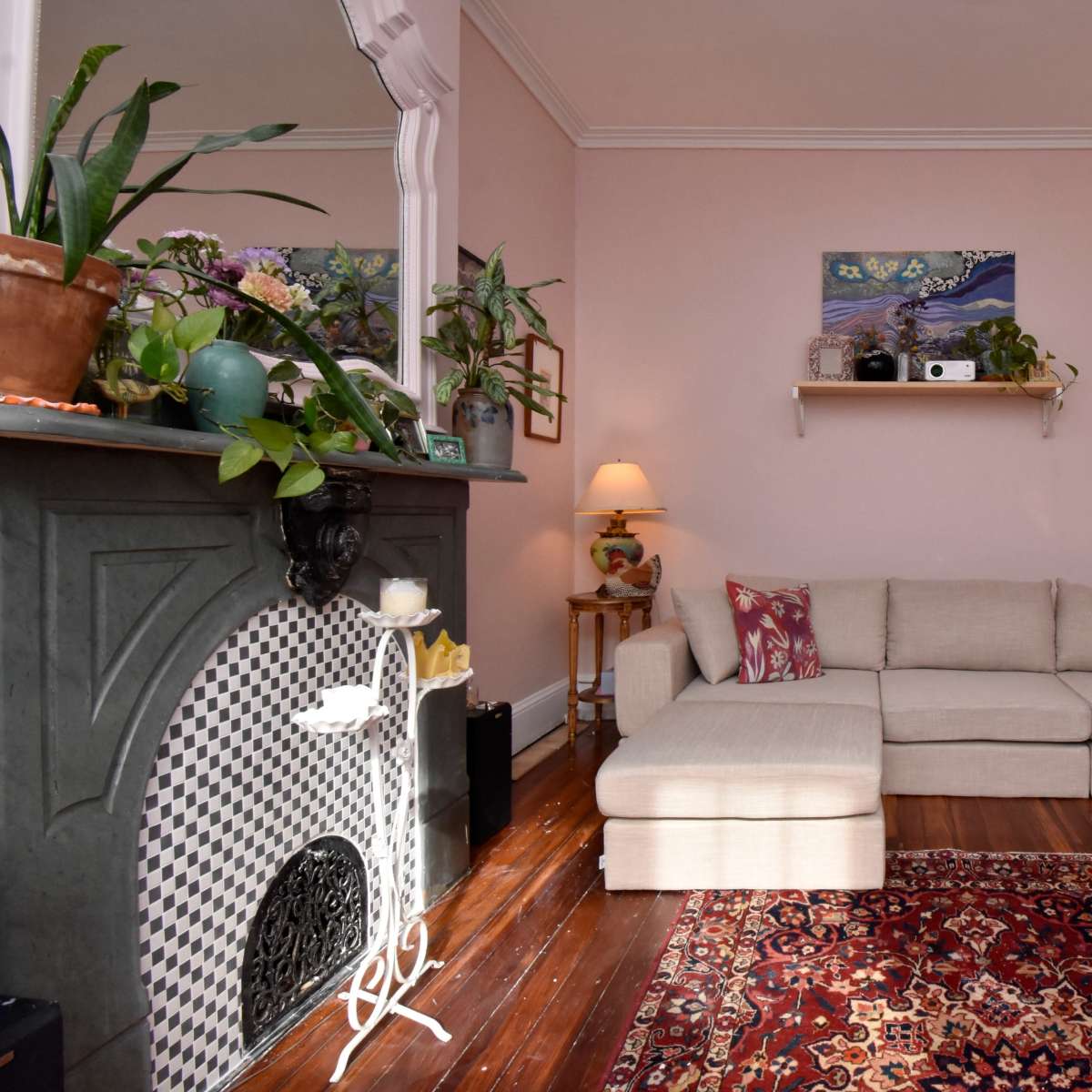 A living room with a light pink wall, a sectional sofa, a patterned rug, and decorative plants on shelves and furniture.