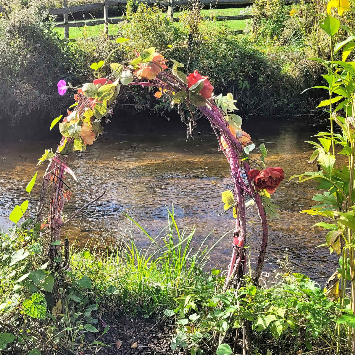A vine-wrapped archway is positioned near a calm stream, surrounded by greenery.