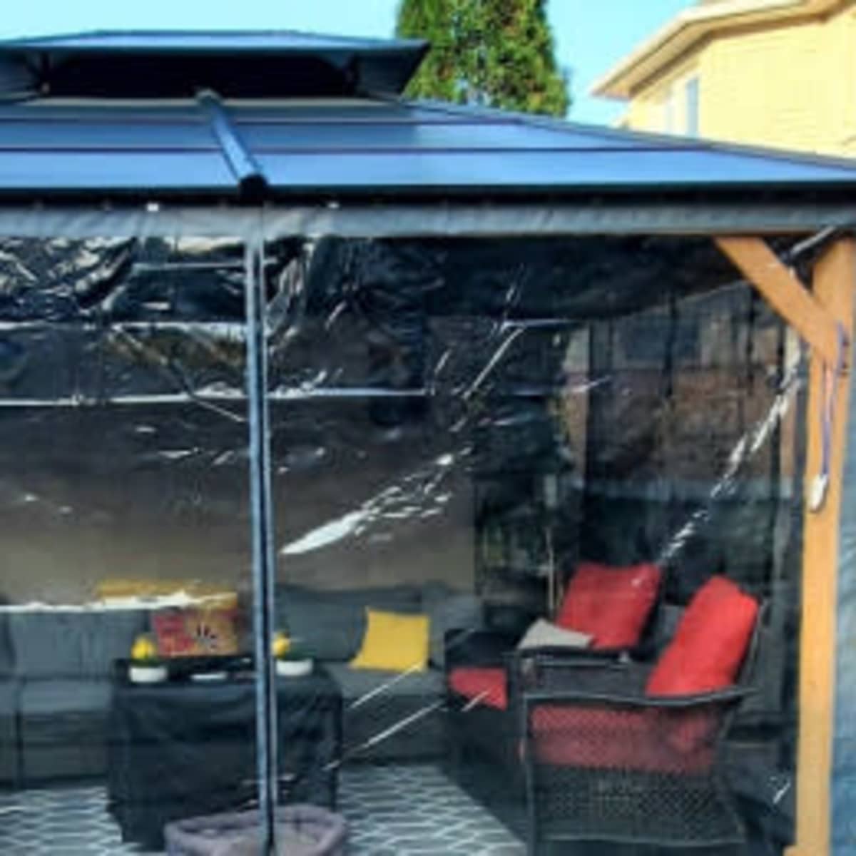 A gazebo with transparent side panels, containing a seating area with red and yellow cushions.