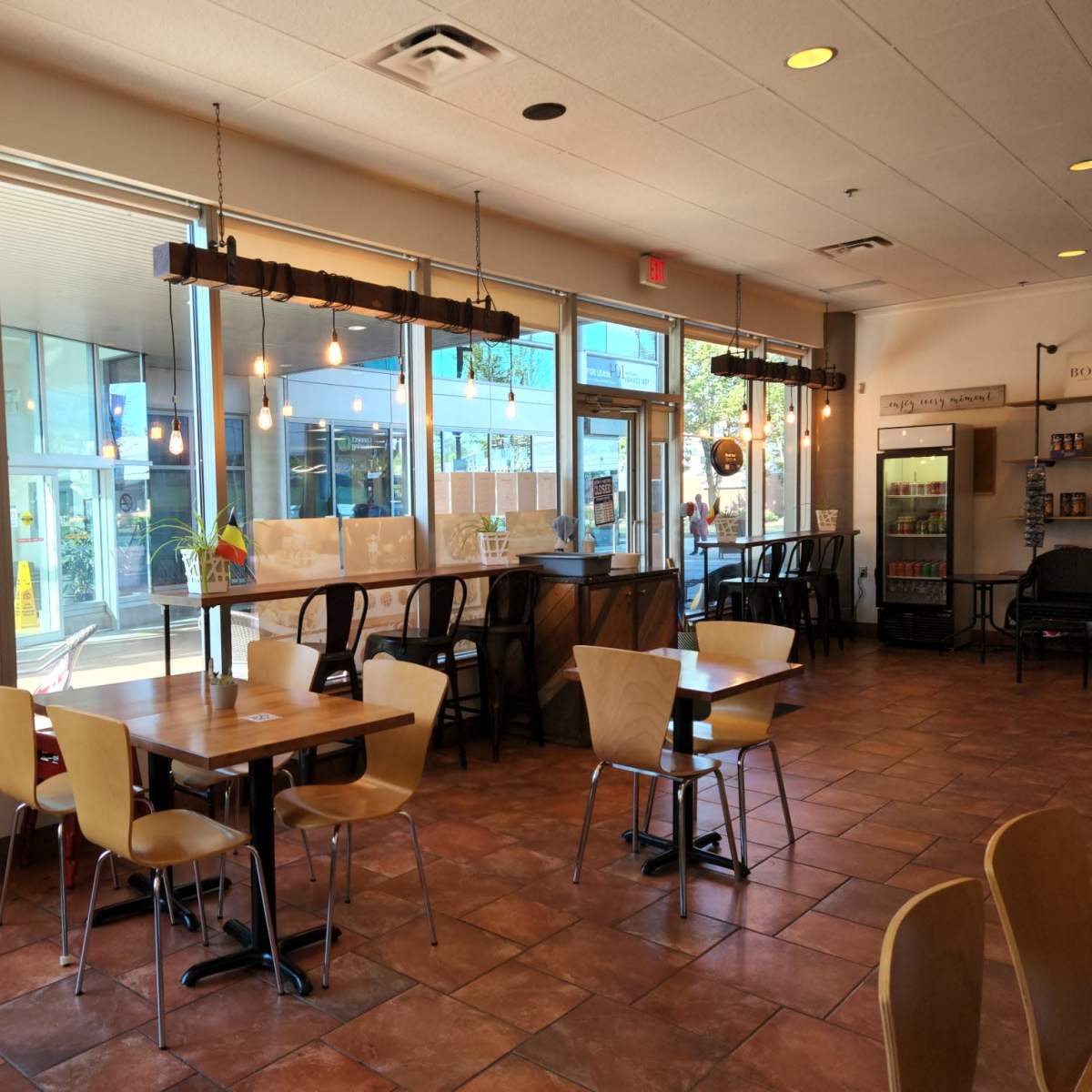 A café interior with tables and chairs, large windows letting in natural light, and a bar counter with hanging lights.