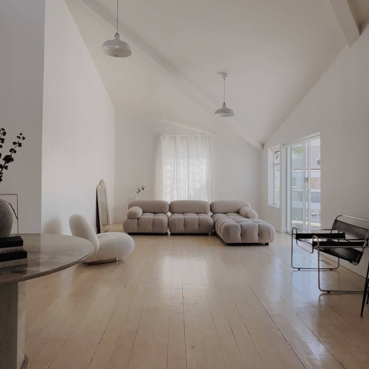A minimalist living room with a light-colored sofa, a round table, and large windows allowing natural light to enter.