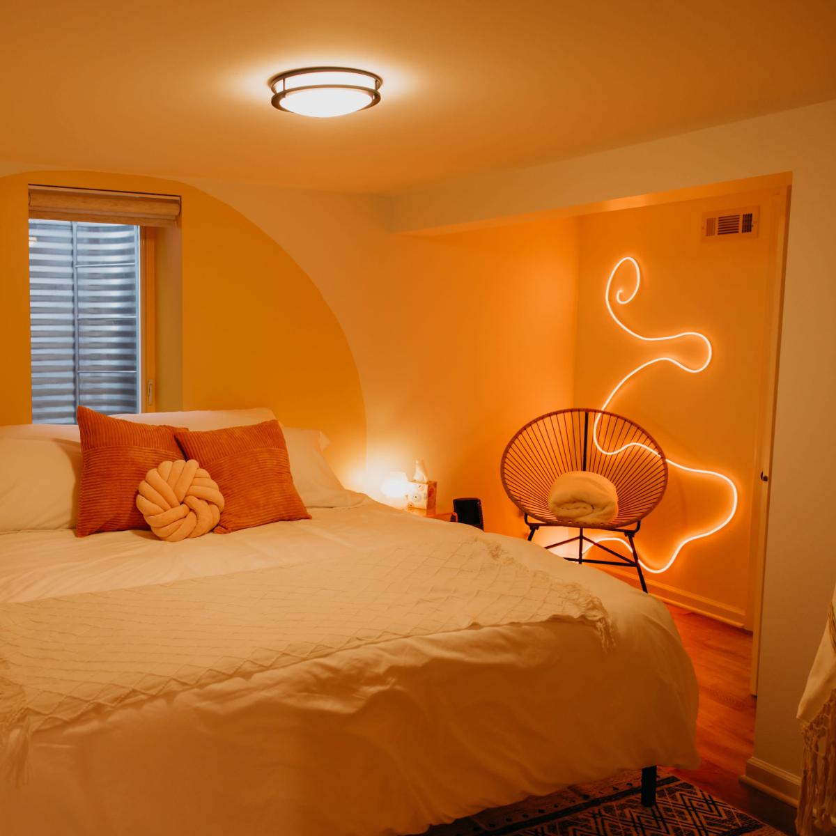 The bedroom features a light yellow accent wall, a neatly made bed with orange pillows, and warm lighting from bedside lamps and a neon design.