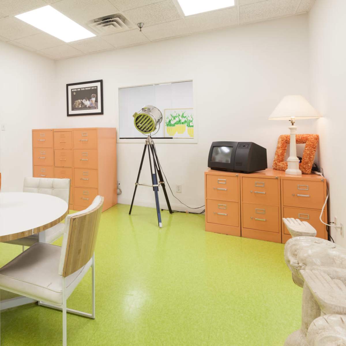 A small office space features a round white table with four chairs, orange filing cabinets, a vintage television, and a large lamp beside a window.