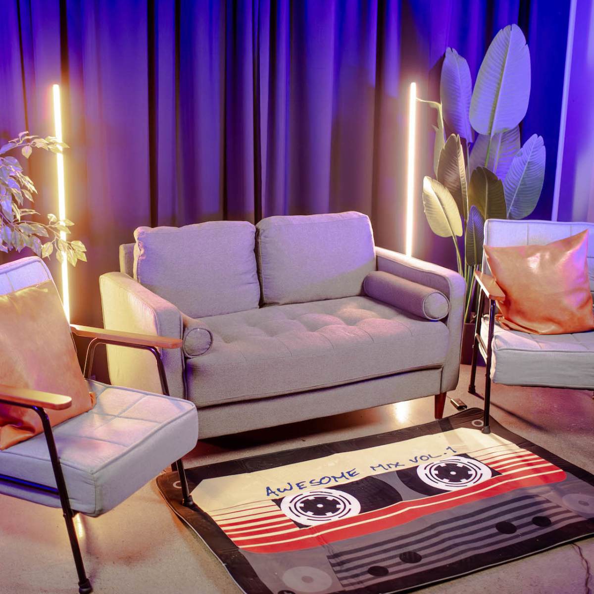 A gray couch with two armchairs and decorative pillows is positioned in front of a rug featuring a cassette tape, all set against a backdrop of blue curtains and illuminated by neon lights.