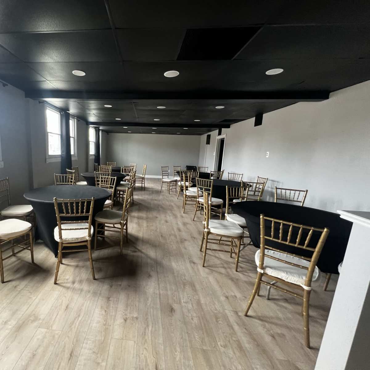 A large event space features several round tables covered with black tablecloths and surrounded by gold chiavari chairs, with windows allowing natural light.